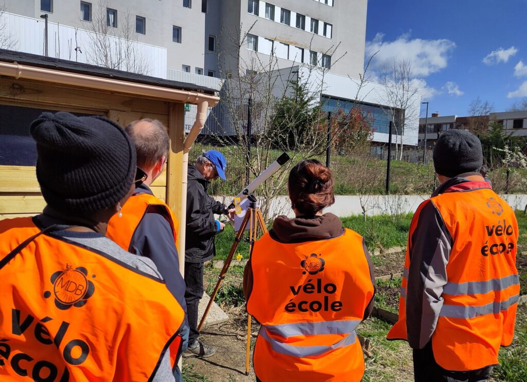 3 membres de la vélo école de dos en train d'observer de loin une lunette astronomique reflétant le soleil sur une feuille de papier