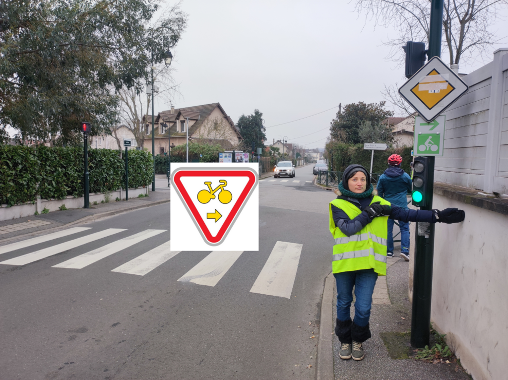 Au pied d'un feu rouge, une cycliste avec un gilet jaune indique avec son bras tendu la droite. Sur sa gauche un panneau M12 Droite